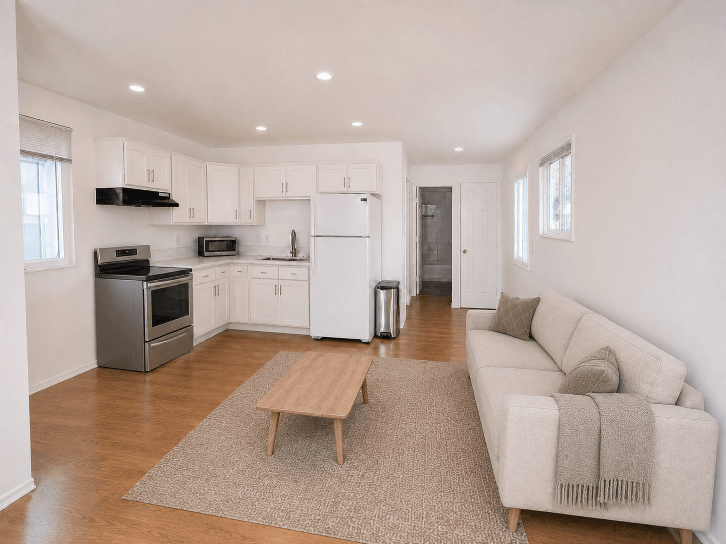 Bright studio apartment with white kitchen cabinets, a stainless steel stove, and a beige sofa.