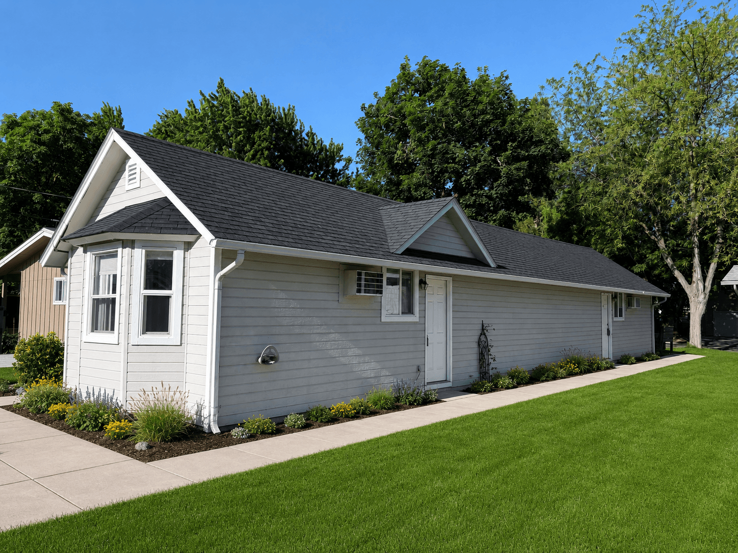 Small beige house with black roof, white trim, and flowers bordering a green lawn.
