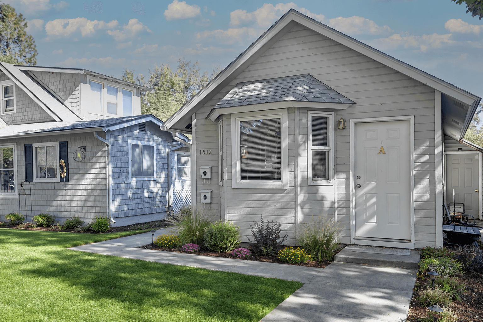 Small gray house with a bay window, white door, concrete path, and green lawn.