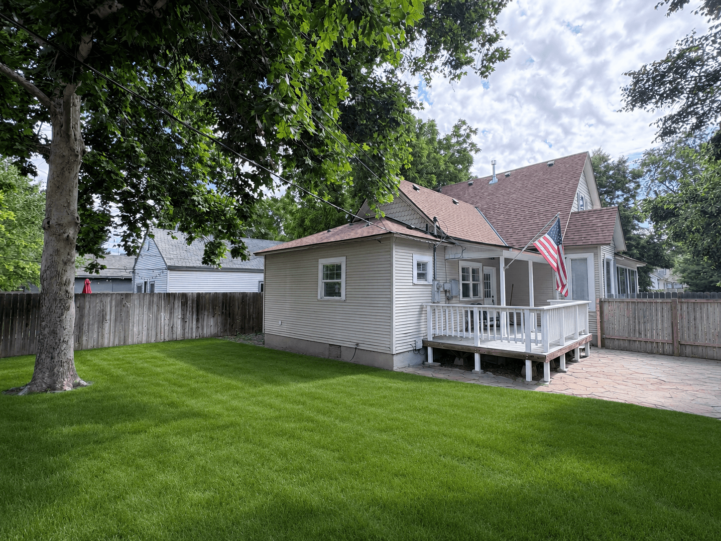 Green backyard with a large tree and house featuring a white deck and American flag.