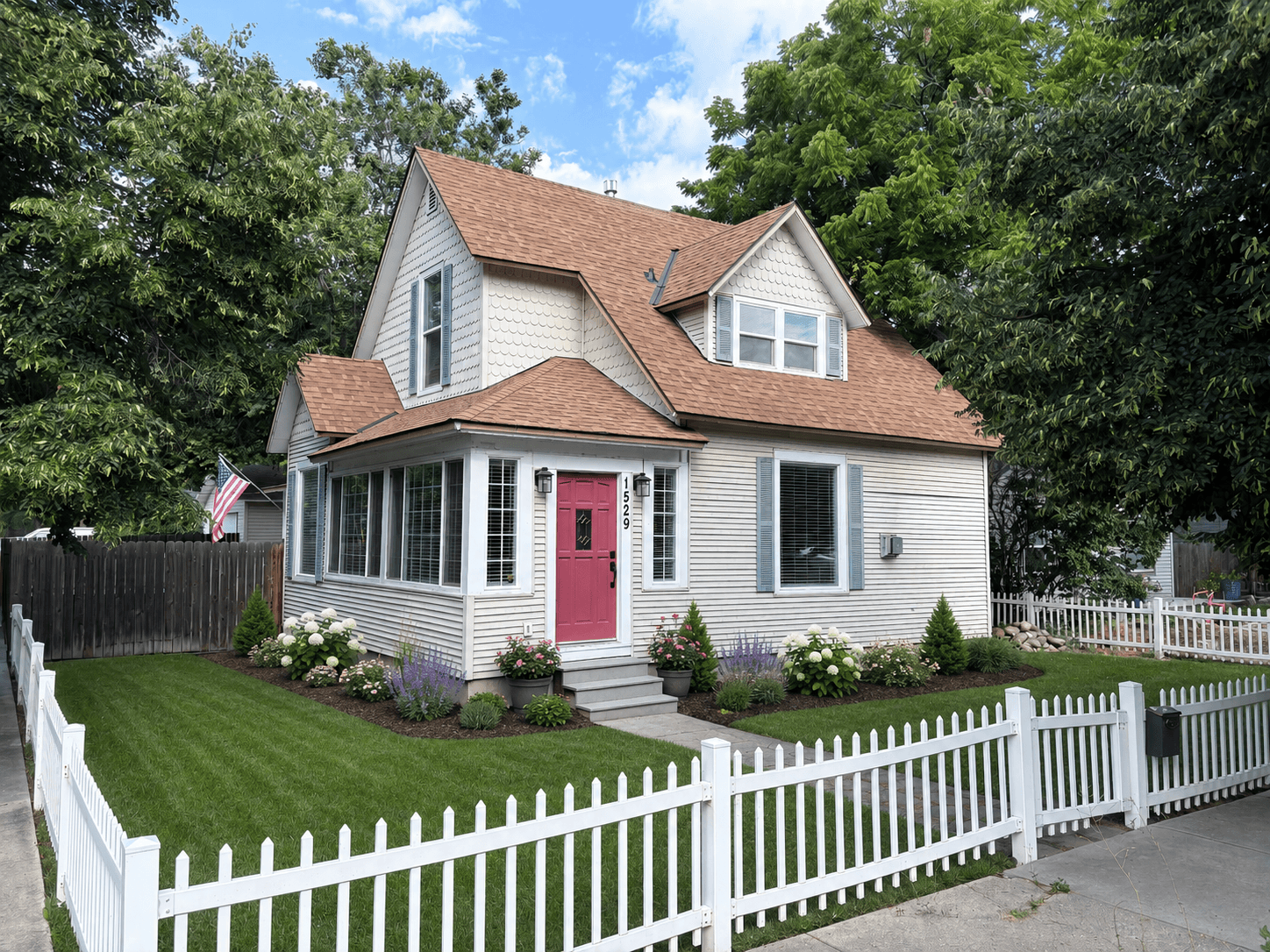 White two-story house with a pink door, white picket fence, and lush green landscaping.