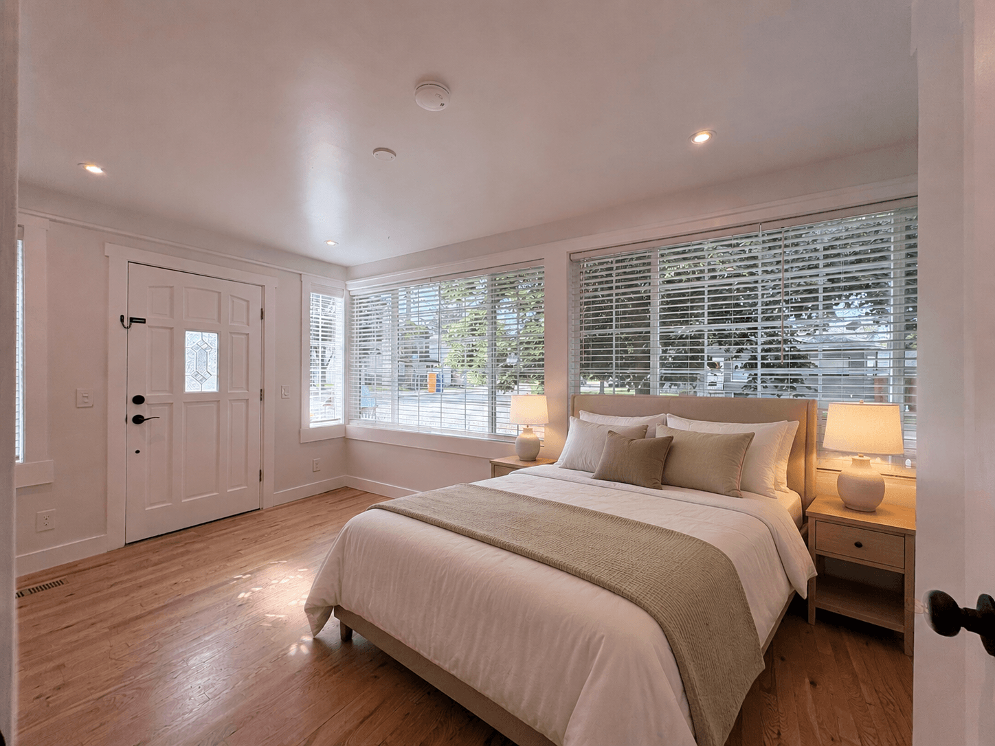 Bright bedroom with a bed, sage green accents, large windows with blinds, and hardwood floors.